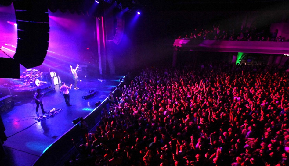 Inside of Hard Rock Live venue in the middle of a concert. Band plays on stage as purple, pink, and blue spotlights shine on them while the packed crowed is lit in red. Orlando, Florida.