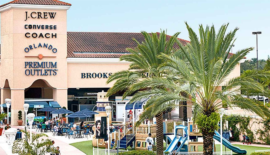Daytime exterior photo of Orlando Vineland Premium Outlets with playground, Brooks Brother's store, and Mall name on building with J-Crew, Converse, and Coach's store names branded above. Shoppers walk the mall. Orlando, Florida.