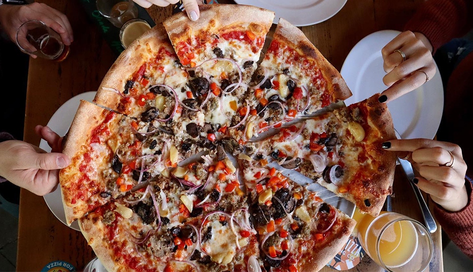 An aerial shot of a full sized pizza (sliced) being taken from at the crust by different people's hands. A drink in a glass sits at the lower right of the photo. Park Pizza & Brewing Co. Lake Nona, Florida. Indoor Photo.