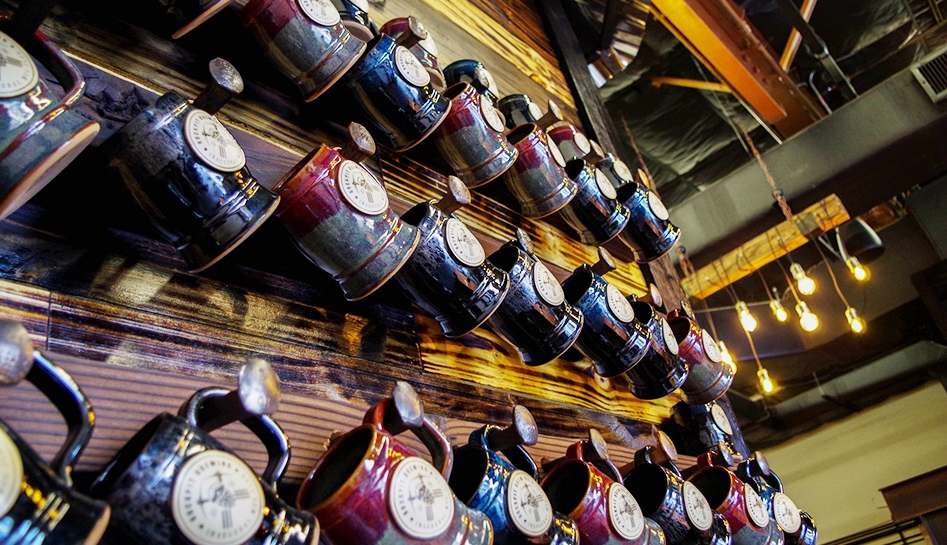 Three rows of mug jugs hanging on a wooden wall with Rockpit Brewing logo printed on each one. Rockpit Brewing. Indoor photo. Orlando, Florida.