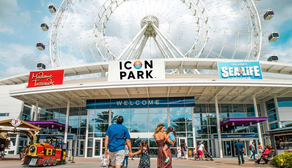 A family of four (from behind from left to right) dad, daughter, and mom holding other daughter as they walk into ICON Park with shot of ferris wheel in the background. Daytime photo. Orlando, Florida.