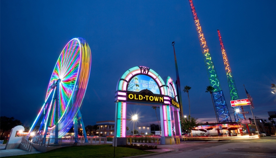 Evening photo of Old Town jukebox entryway design (center) ferris wheel lit up (left) and slingshot attraction (right). Kissimmee, Florida.
