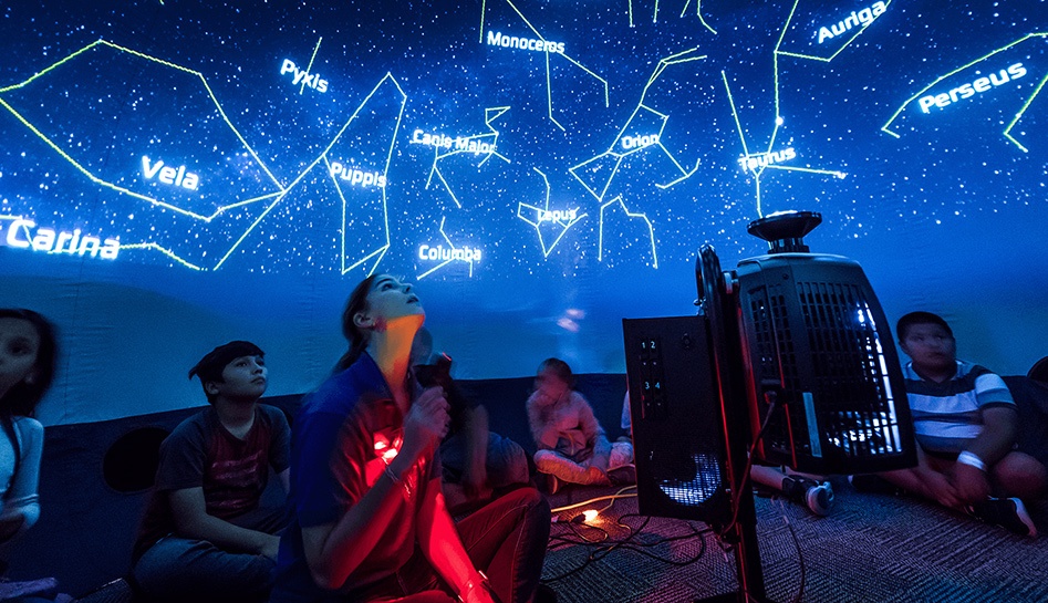 A woman and multiple children sit on the floor as they look up at the constellations with names lit up in blue and white on the ceiling at Orlando Science Center. Orlando, Florida.