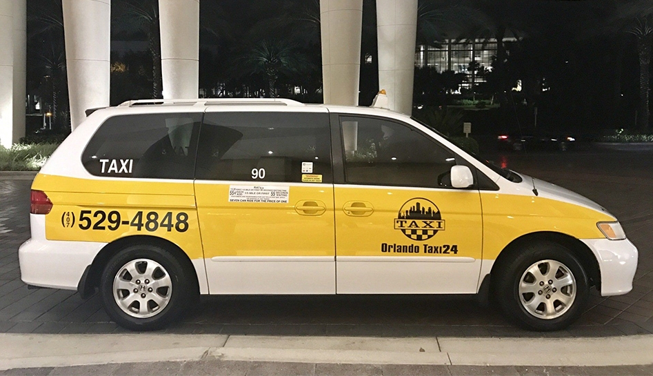 Nighttime photo of passenger side of white and yellow taxi van with company logo on the front passenger door. Orlando Taxi 24. Orlando, Florida.