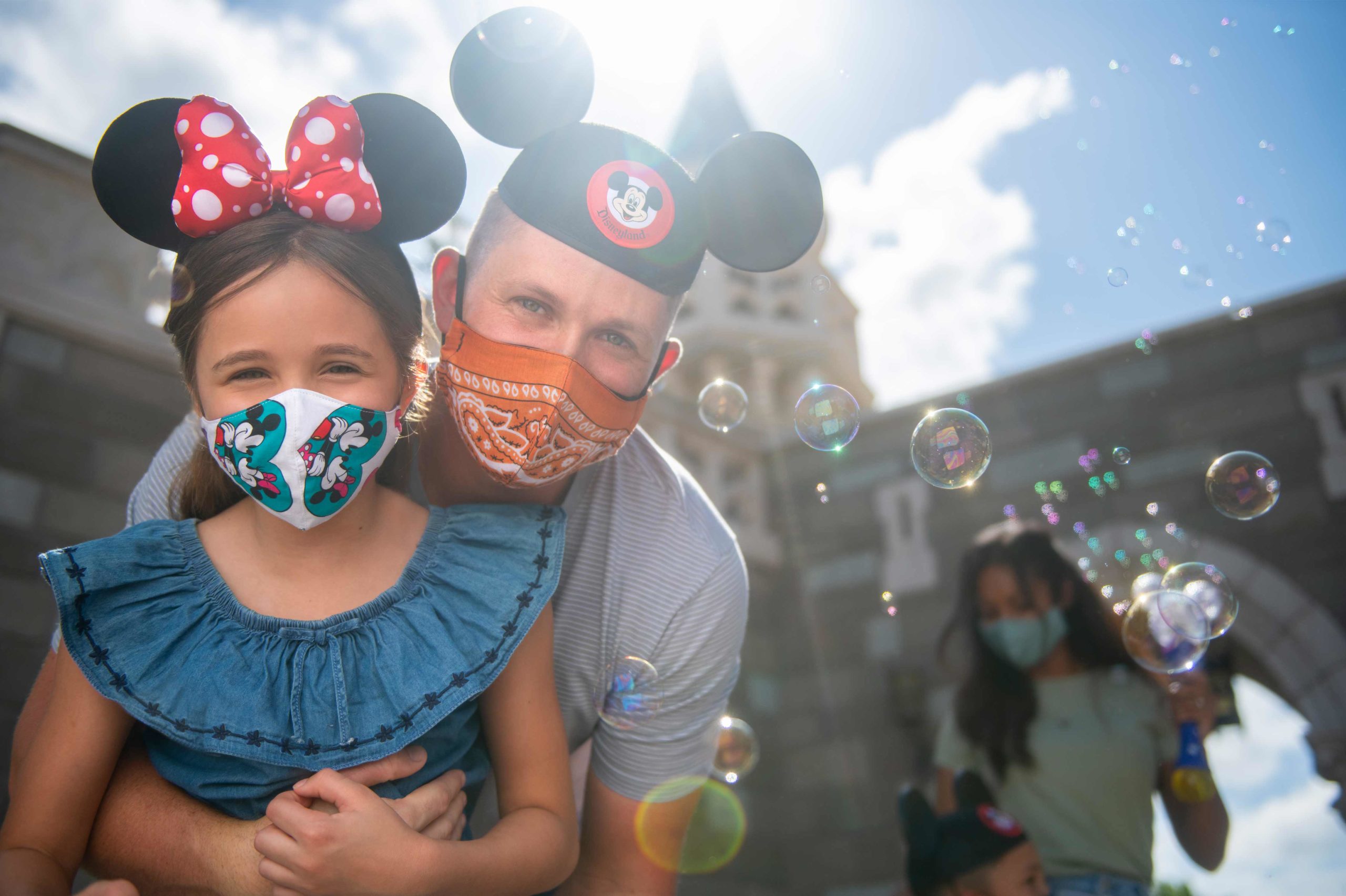 Father giving daughter a back hug for a photo while wearing face masks near the Cinderella Castle at the Magic Kingdom® Park. Daytime shot. Walt Disney World® Resort. Lake Buena Vista, Florida. ©Disney.