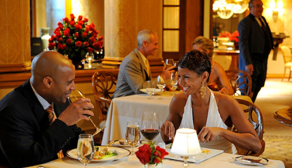 Male (left) and female (right) couple smile at one another as they sit at the dining table with dinner at Victoria & Albert's. Guests and waiter also in background. Disney's Grand Floridian Resort & Spa. Walt Disney World® Resort. Orlando, Florida.