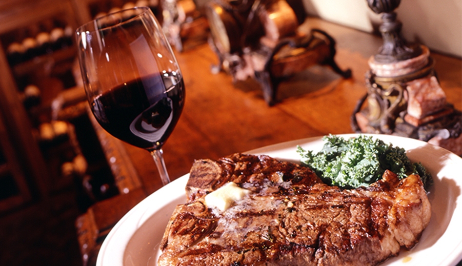 Angled photo of a glass of red wine (left) and an entree of steak with leafy greens (right) on a wooden table inside of Vito's Chop House. Orlando, Florida.