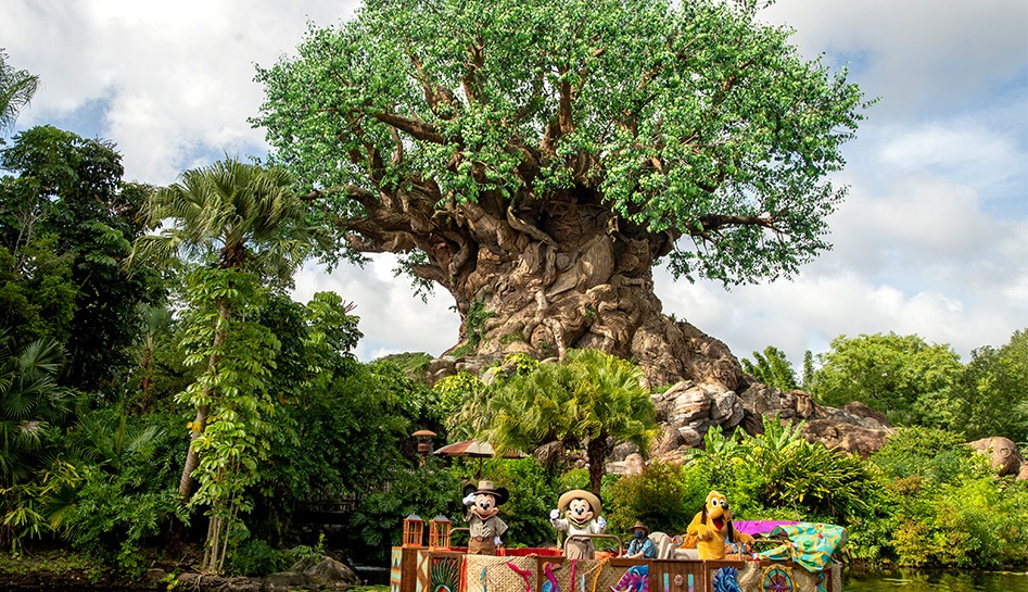 Daytime photo of the Tree of Life with a boat sailing in the river in front. On the bote, you can see Mickey Mouse (left), Minnie Mouse (middle), and Pluto (right). Mickey and Minnie are waving toward the direction of the camera. Disney's Animal Kingdom® Theme Park. Lake Buena Vista, Florida. Orlando.